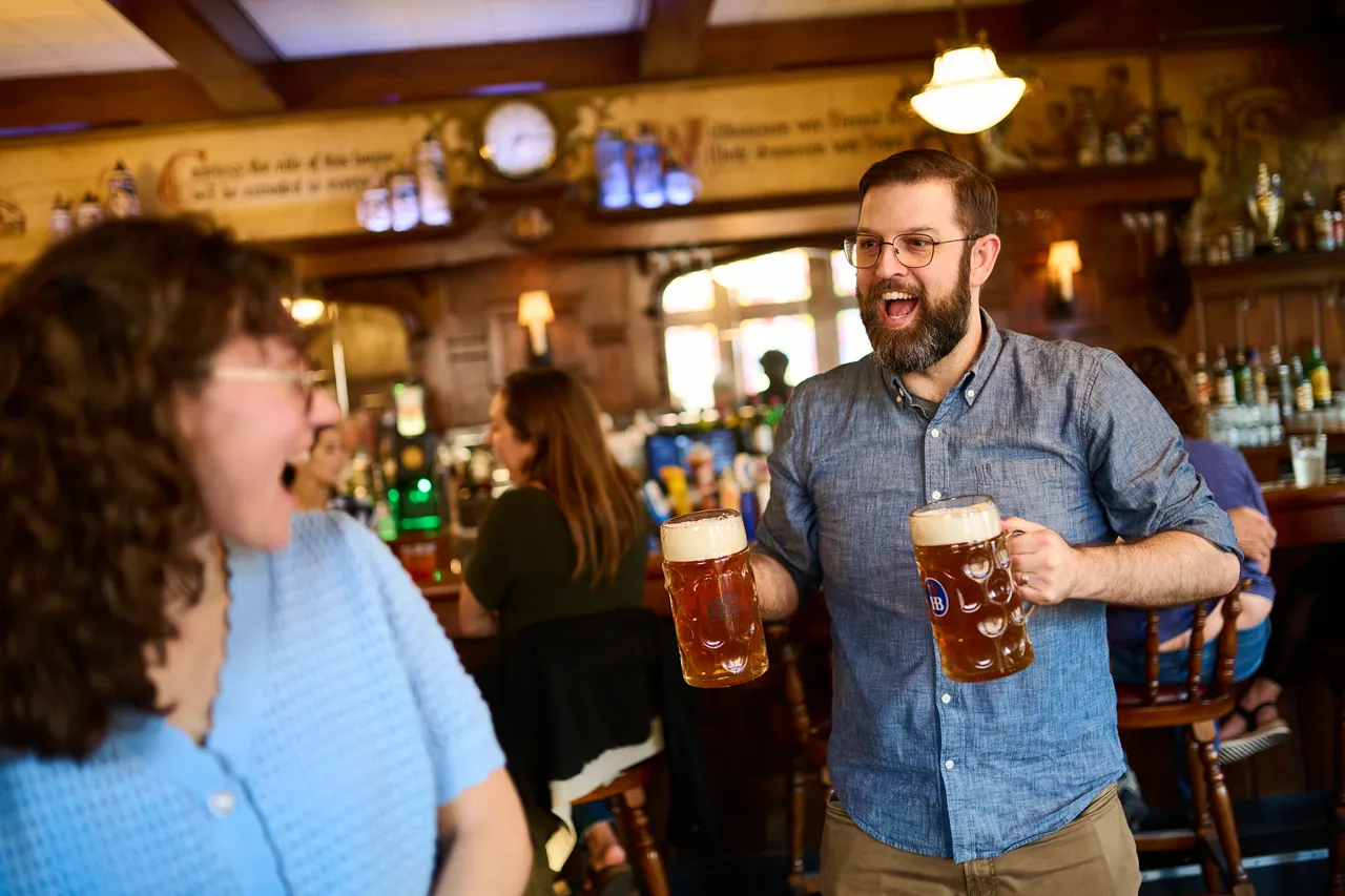 Friends laugh and raise large beer steins inside a warmly lit pub.