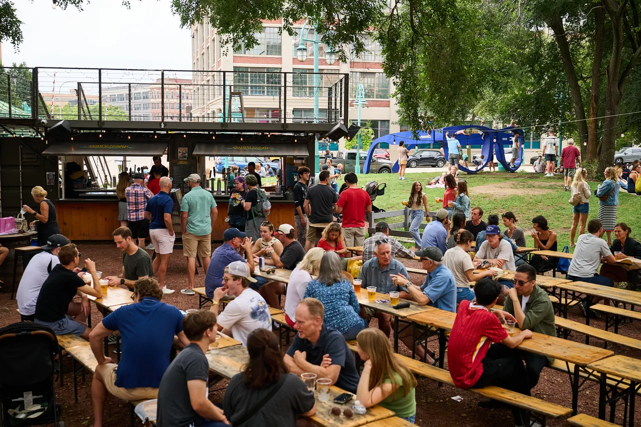 People dine at picnic tables beside a food truck in a leafy park with a blue sculpture nearby.