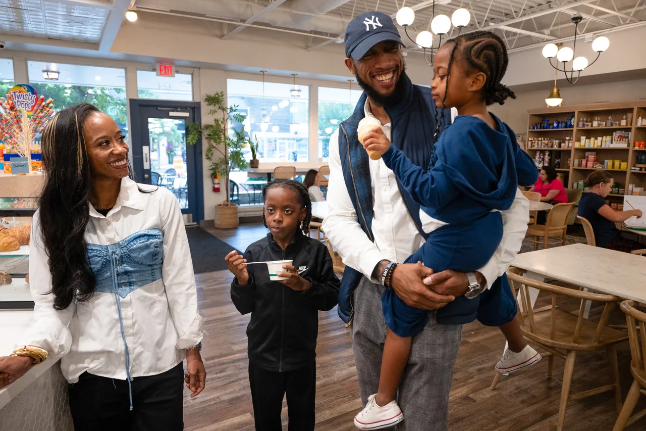 A family shares a meal in a bright café, with a father holding one child as another eats nearby.