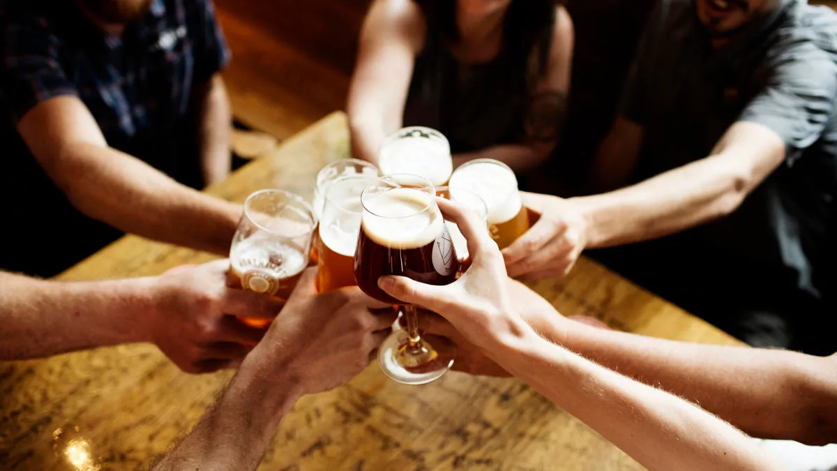 Friends clink glasses of beer over a wooden table in a warmly lit bar.
