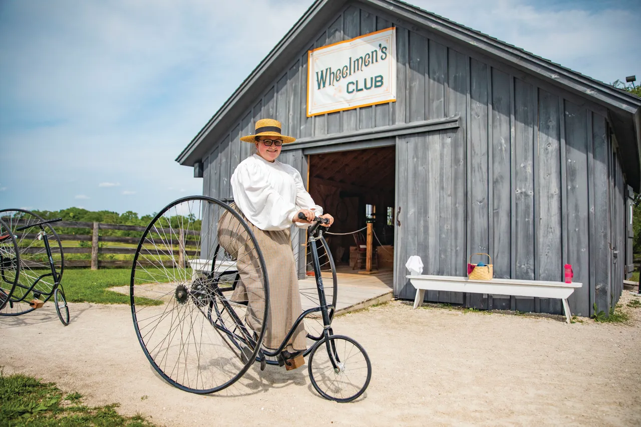 A person in period clothing stands beside a high-wheel bicycle outside a gray Wheeling Club building.