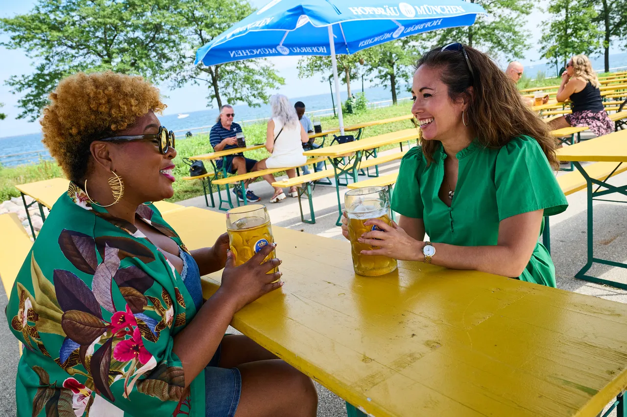 Two women laugh together over drinks at a yellow picnic table beside Lake Michigan.