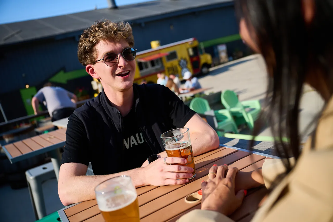 A man wearing sunglasses talks with a friend over drinks at a sunny outdoor beer garden.