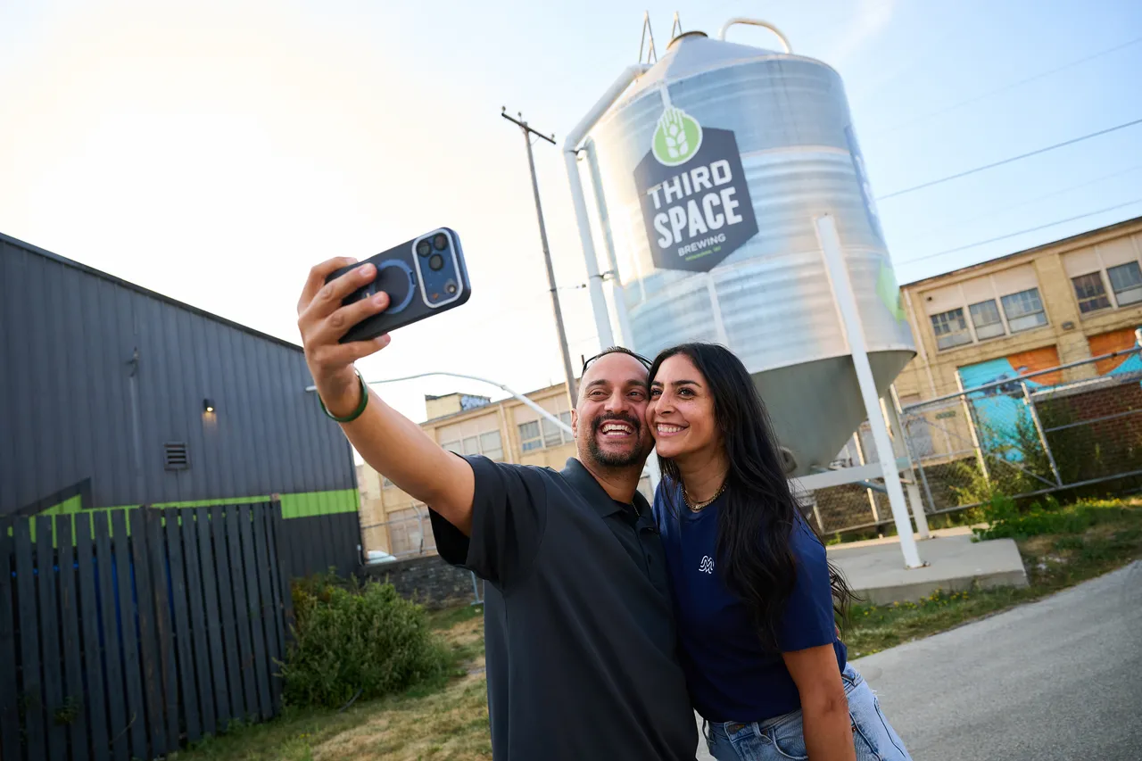 A smiling couple takes a selfie in front of Third Space Brewing’s large silver silo.