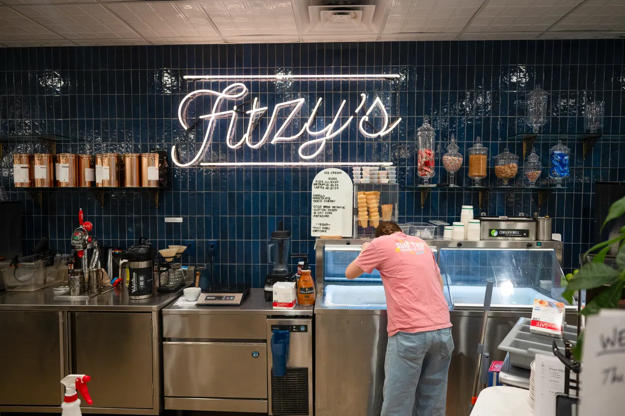 A worker in a pink shirt prepares treats behind the counter beneath Fitzy’s neon sign.