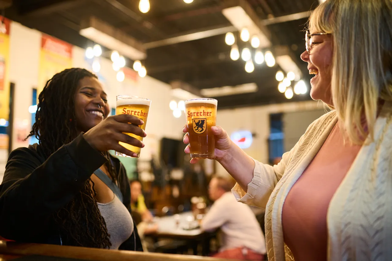Two women smile as they raise glasses of Sprecher beer for a toast in a lively taproom.