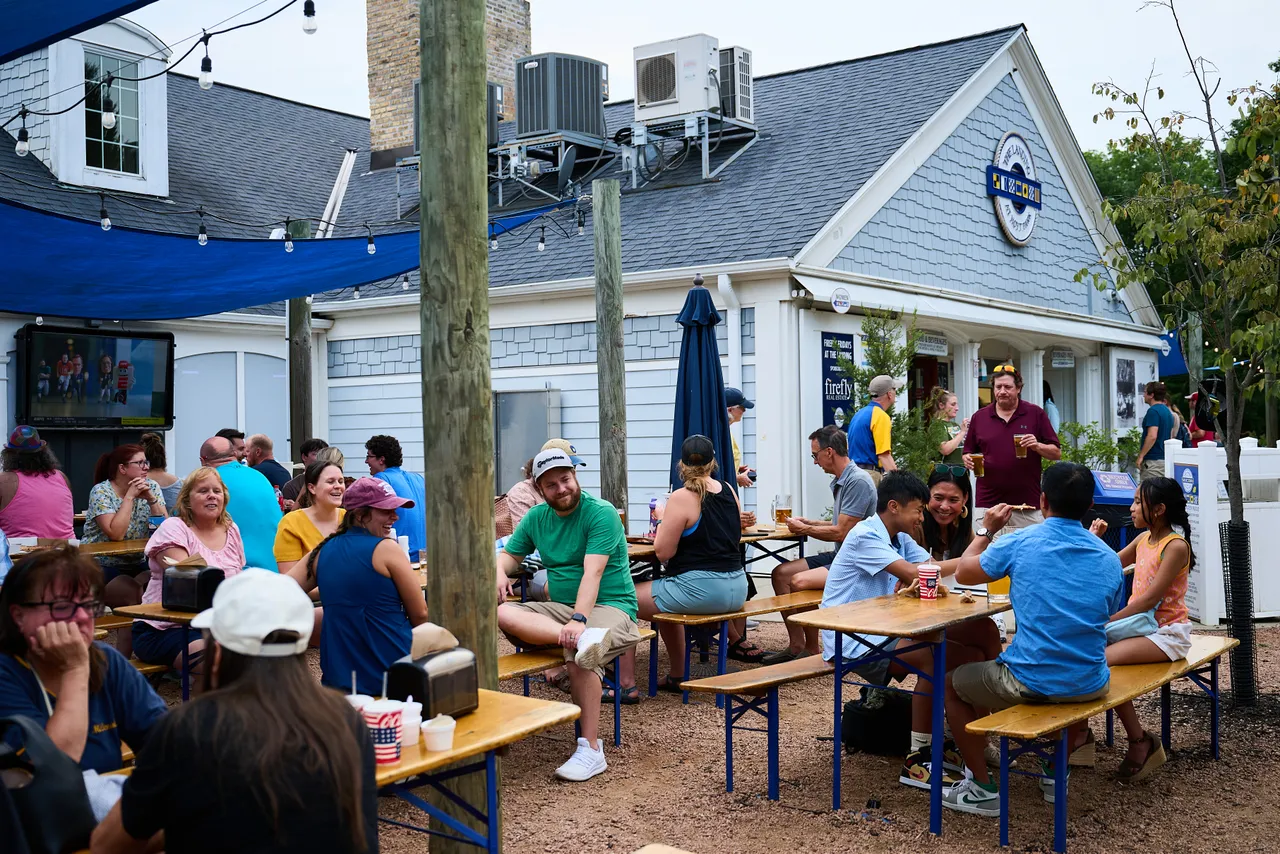 People gather at picnic tables on a lively patio outside a blue-and-white restaurant.
