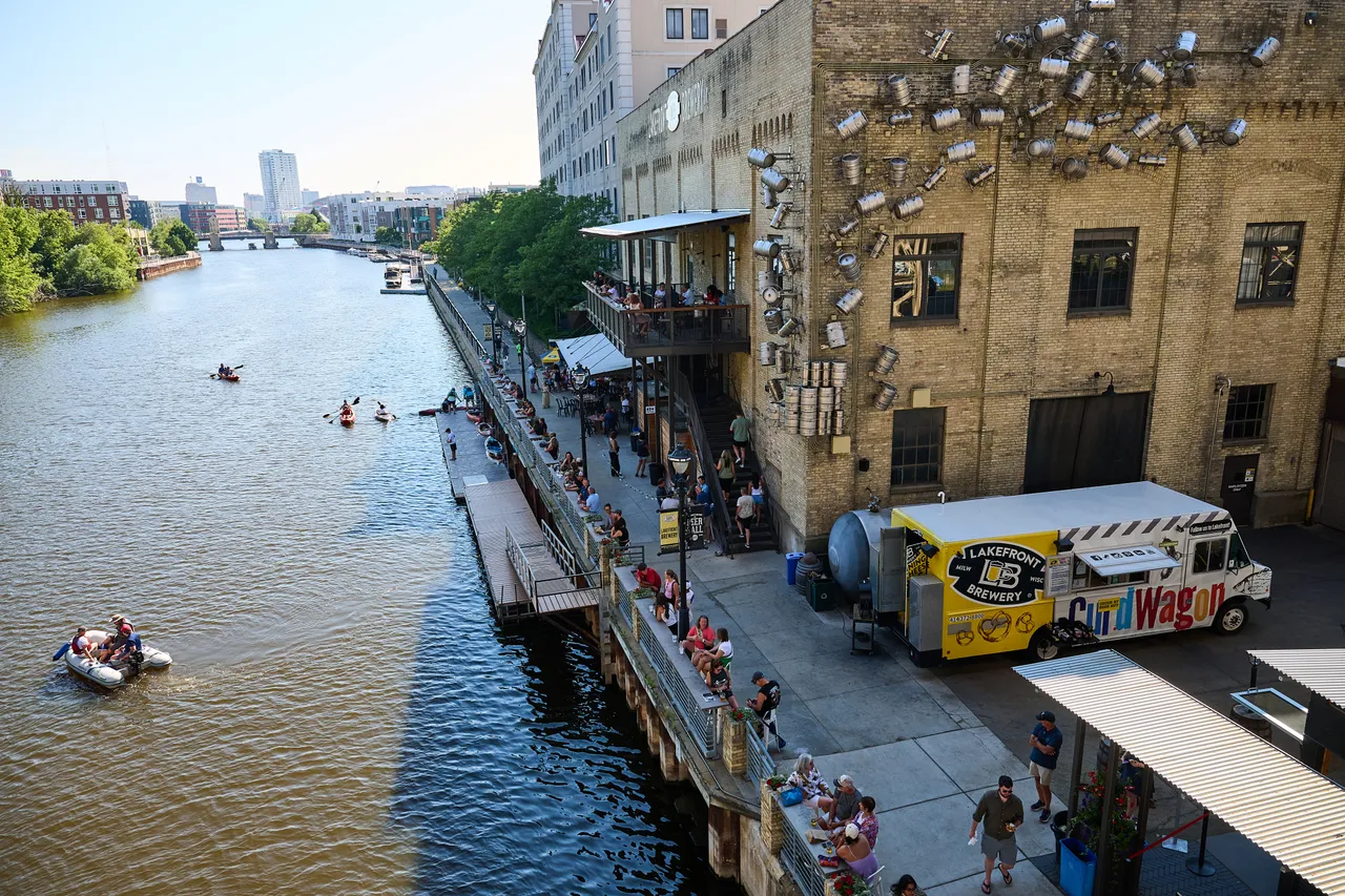Kayakers paddle along the Milwaukee River beside a crowded riverwalk and brick brewery building.