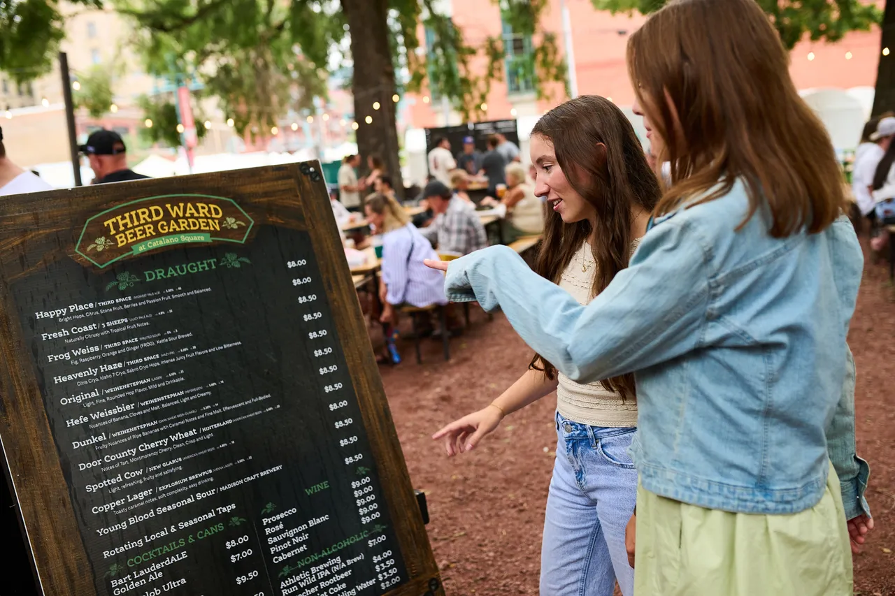 Two women point out drinks on a menu at an outdoor beer garden.