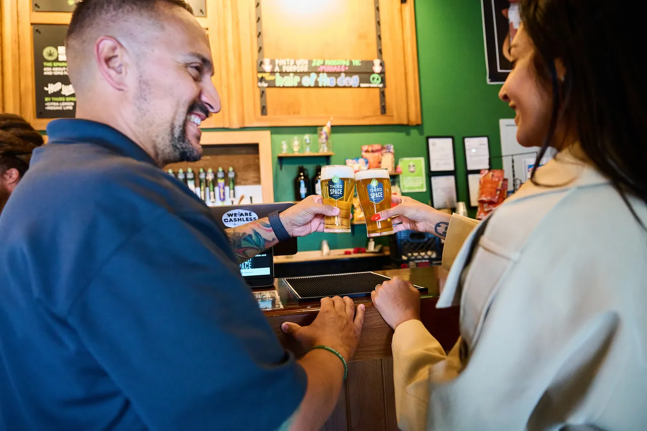 A smiling customer and shop worker exchange canned drinks across a store counter.