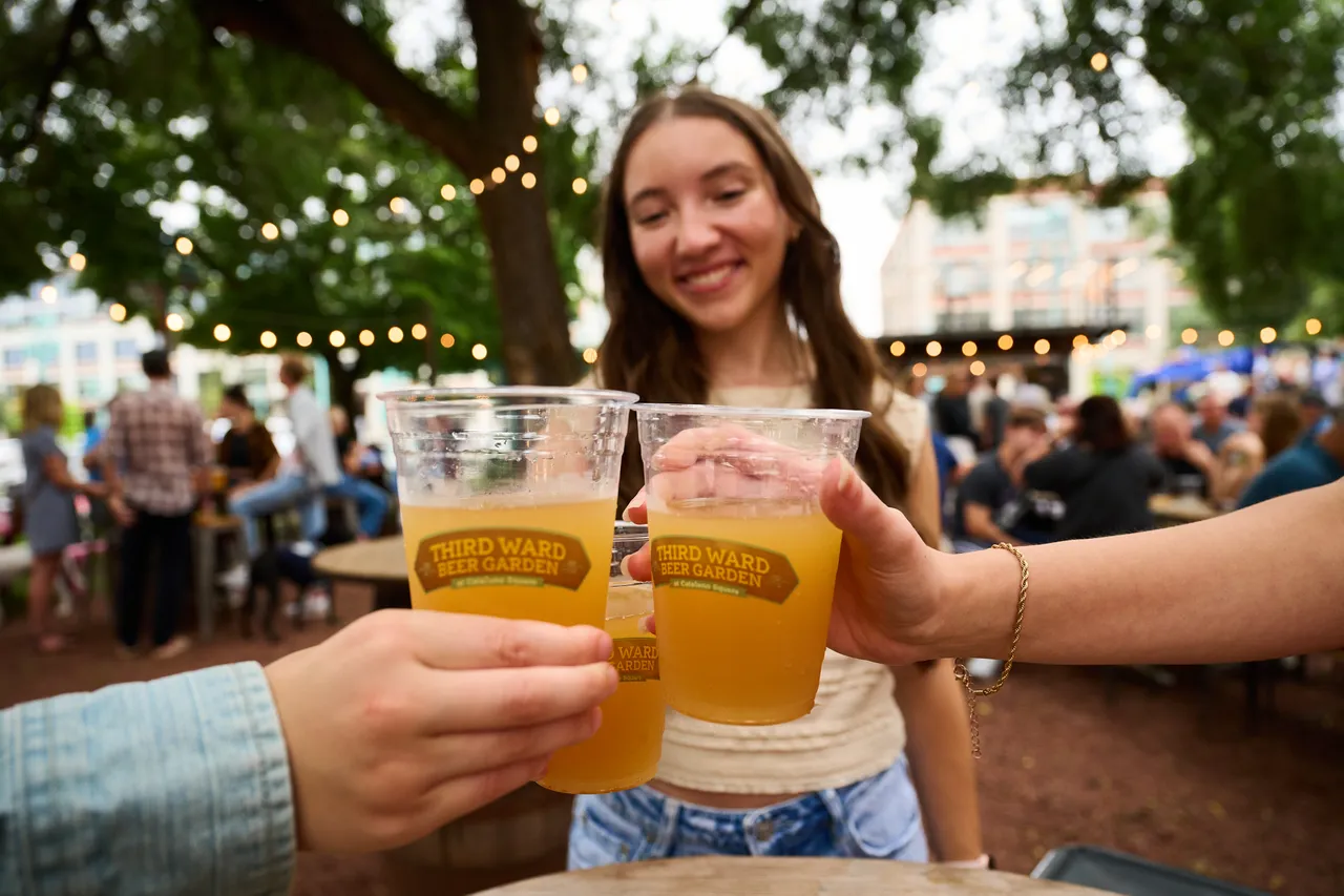 Friends toast with cups of beer at a lively outdoor beer garden under string lights.
