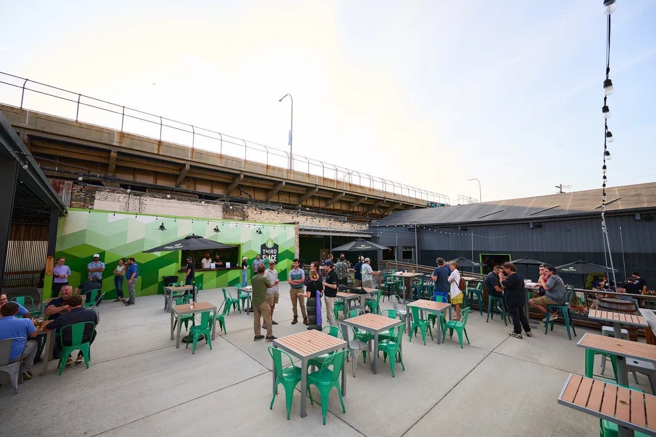 Visitors gather around green tables in an outdoor beer garden beneath an elevated highway.