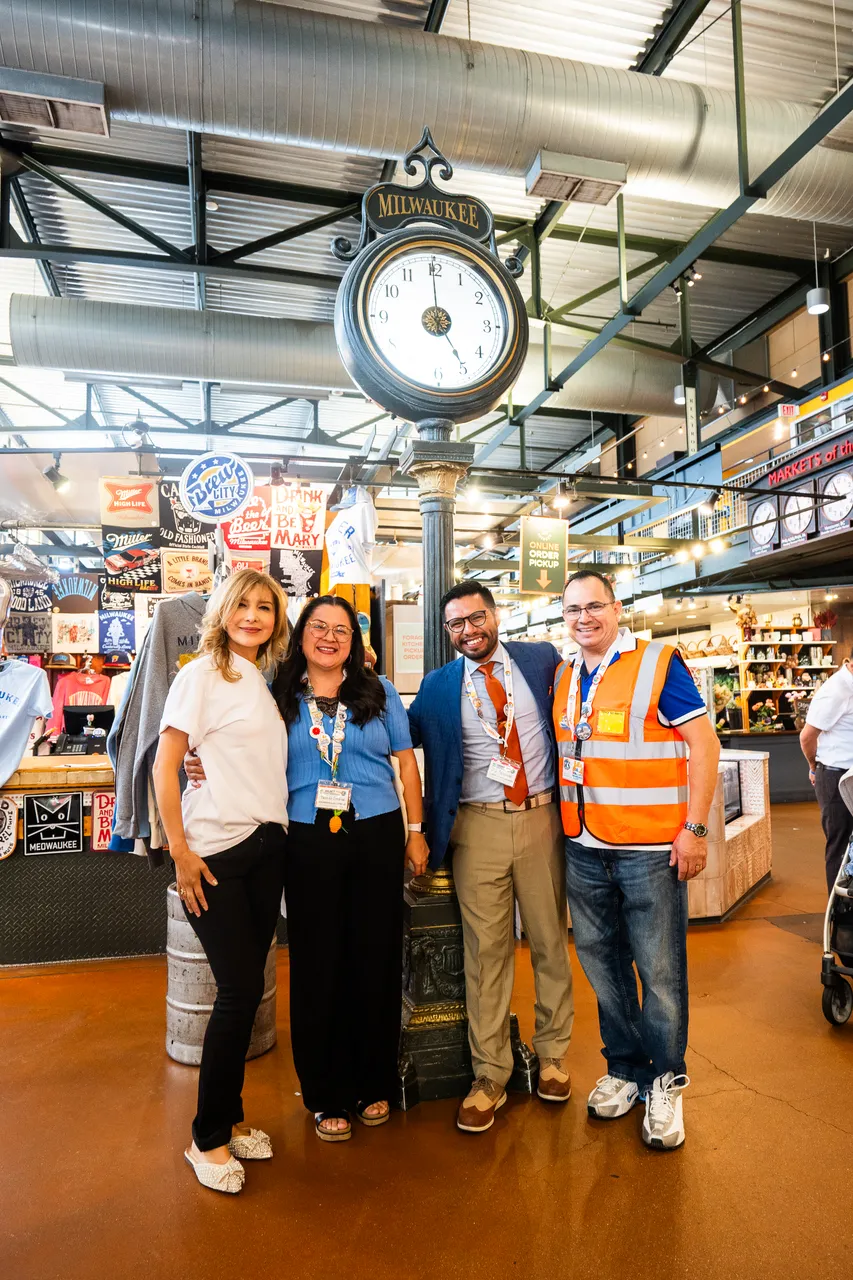 Four people pose beside the Milwaukee clock inside a busy market.