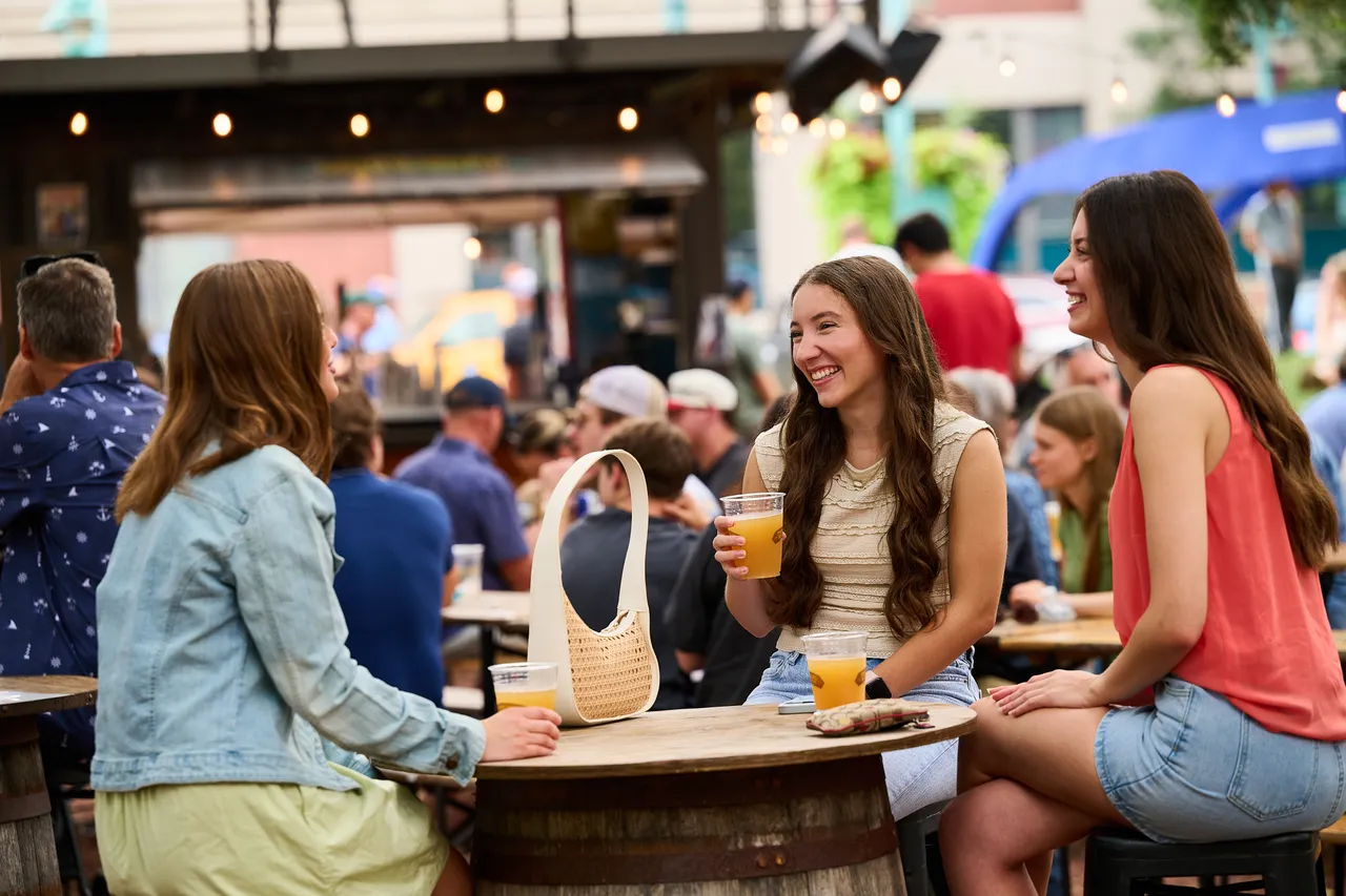 Three friends chat over drinks at a crowded outdoor food hall.