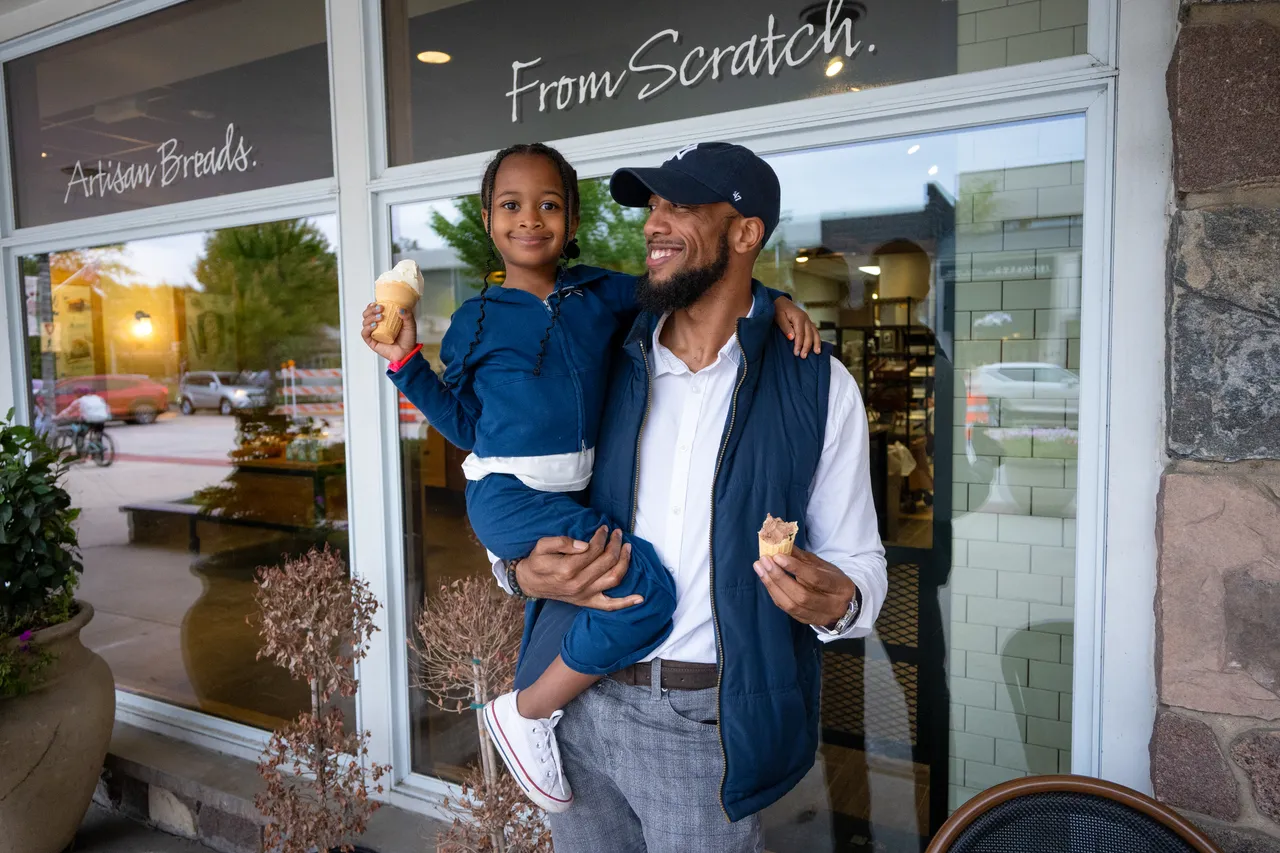 A father holding his daughter smiles outside a bakery while they enjoy ice cream.
