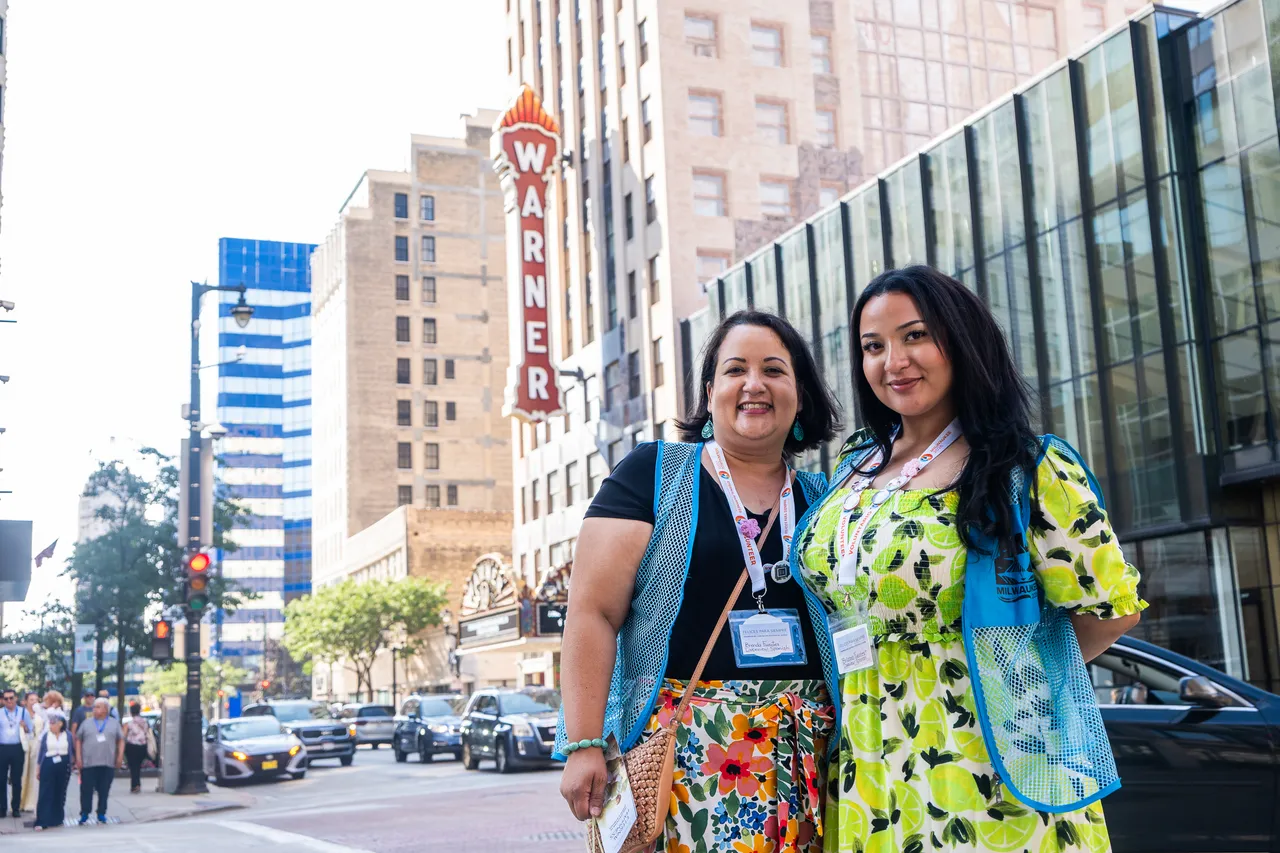 Two smiling women in colorful outfits pose on a downtown Milwaukee sidewalk near the Warner Theatre sign.