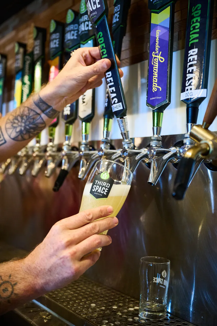 A tattooed bartender pours pale beer from a brewery tap into a glass.