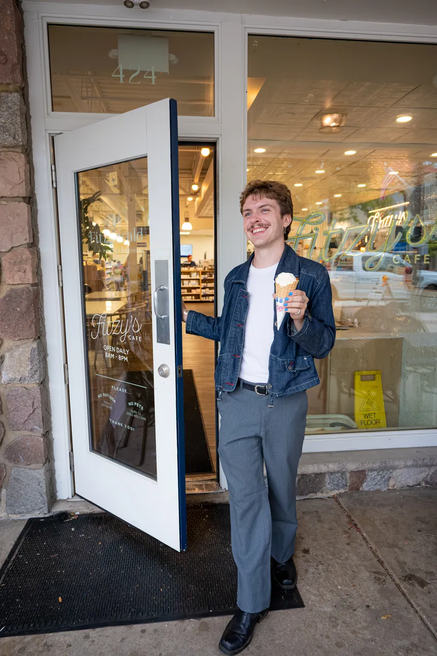 A smiling man holds an ice cream cone in the open doorway of a cafe.