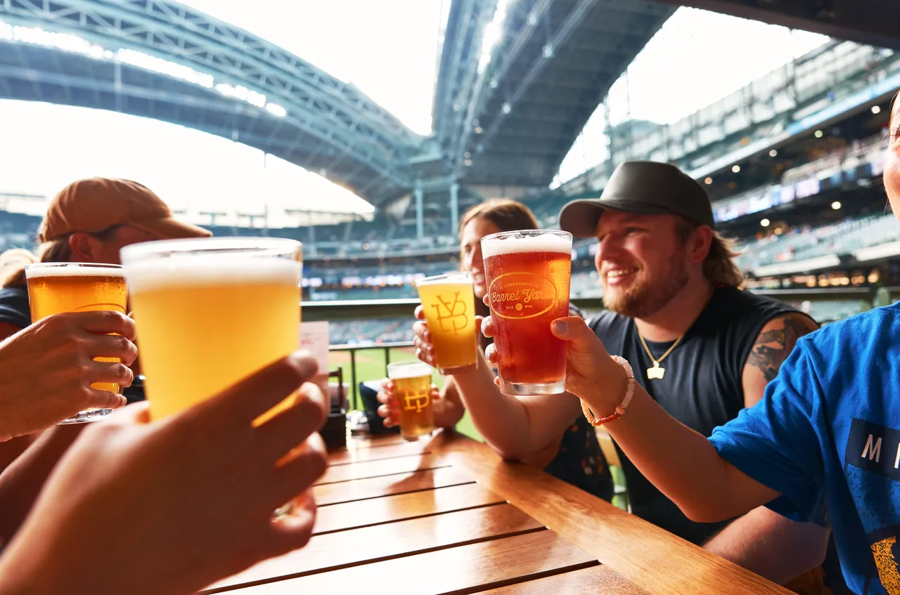 Friends raise glasses of beer around a table at a stadium.