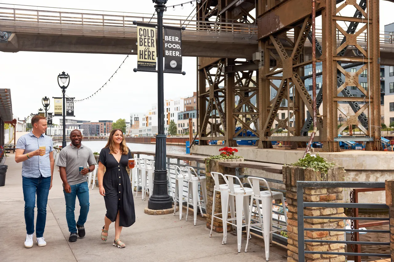 Three friends walk past riverfront patio seating beneath a steel bridge in Milwaukee.
