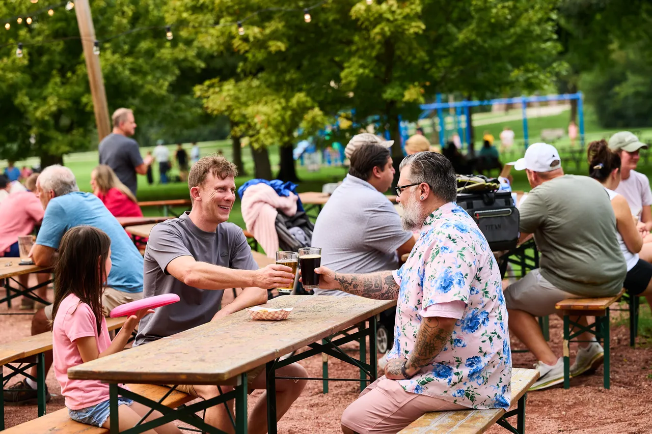 Two friends toast with beer at a busy outdoor beer garden surrounded by picnic tables and trees.