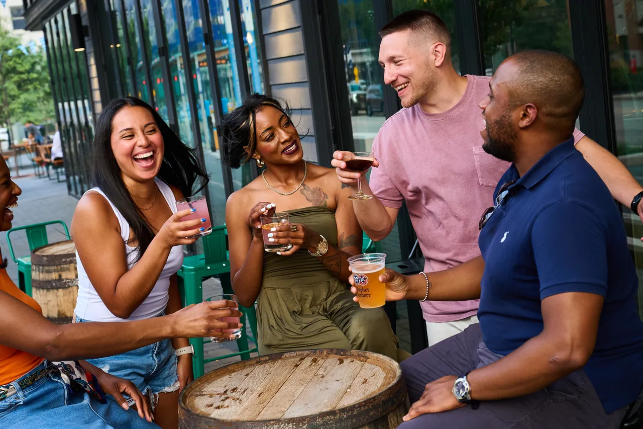 Friends laugh and chat over drinks around a barrel table on an outdoor patio.