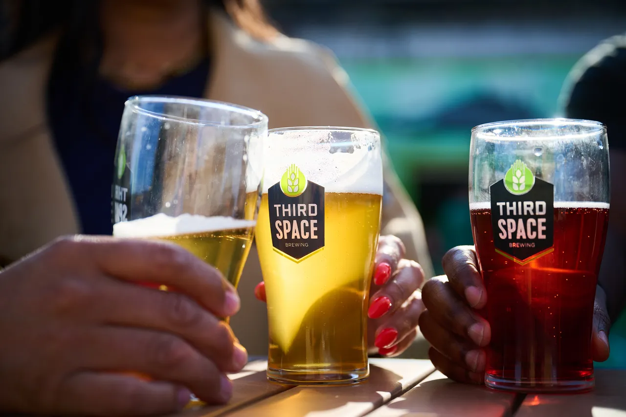Friends hold glasses of golden and red Third Space Brewing beer at an outdoor table.