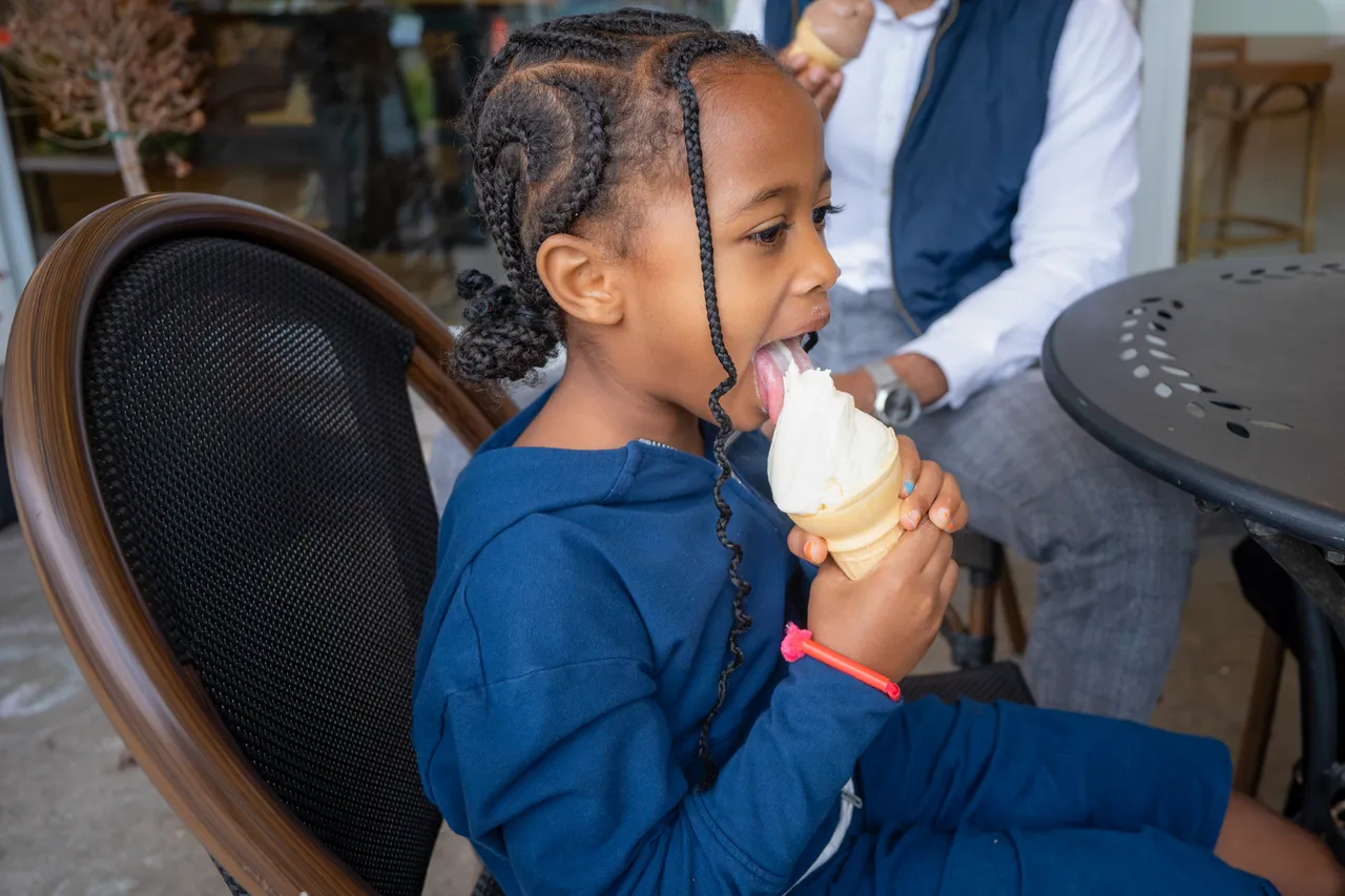 A young girl in blue enjoys a vanilla ice cream cone while seated at a café.