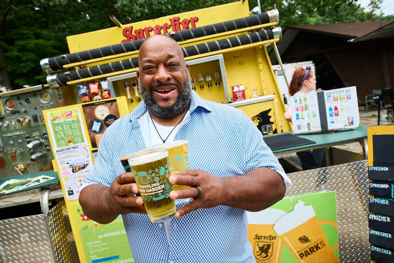 A smiling man holds a beer in front of a yellow beer garden stand.
