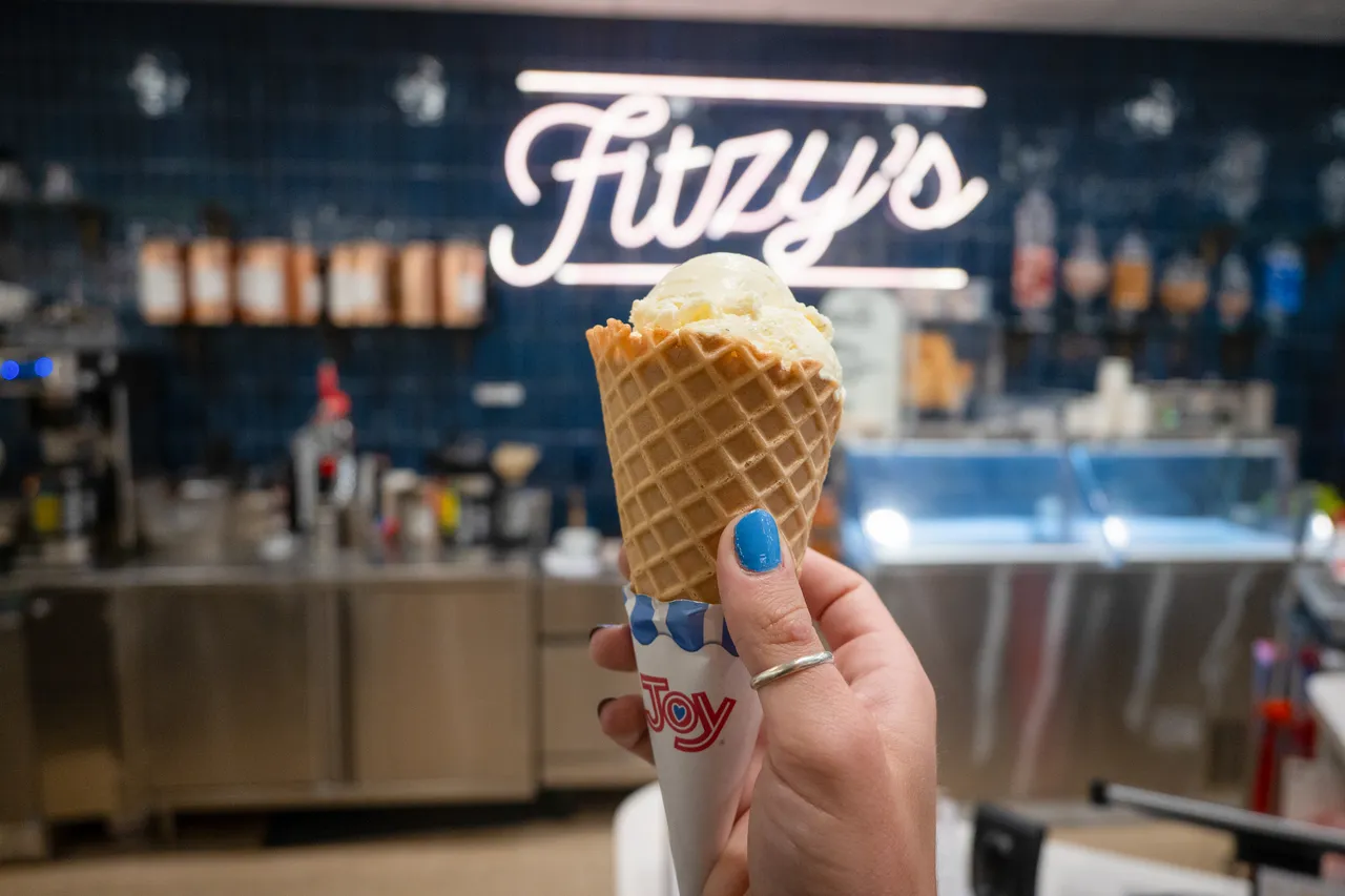 A hand with blue nail polish holds a waffle cone topped with ice cream in front of Fitzzy’s sign.
