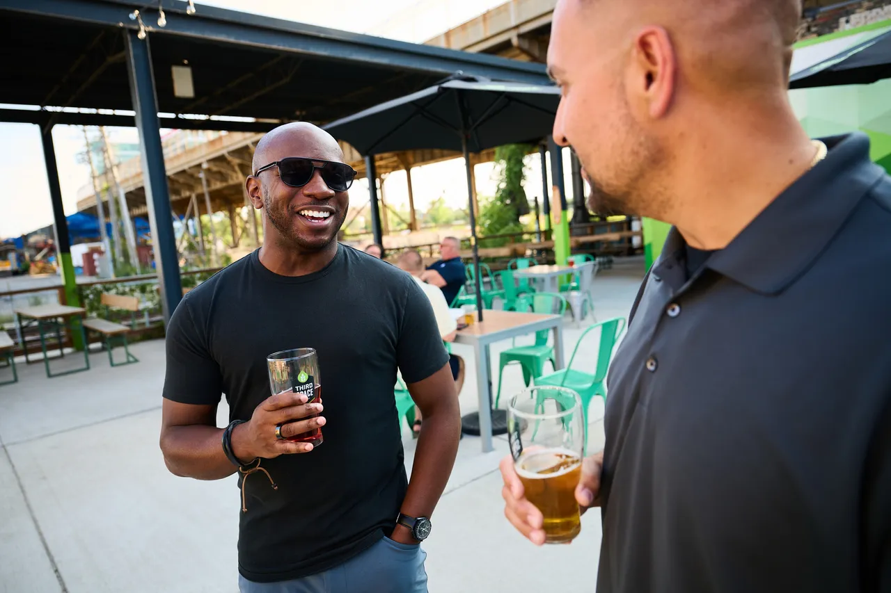 Two men chat over drinks on a sunny outdoor patio with green chairs and tables.