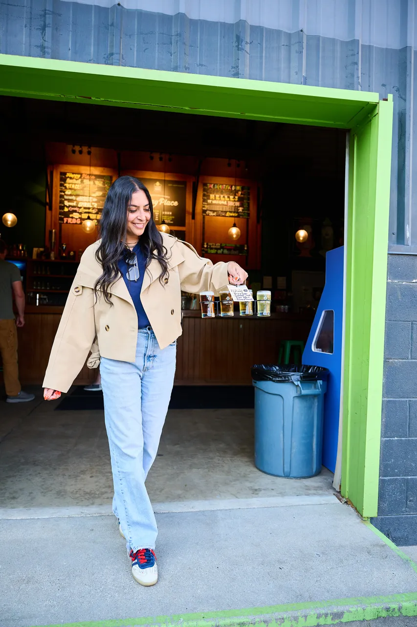 A woman in a beige jacket walks out of a shop with a small item in hand.