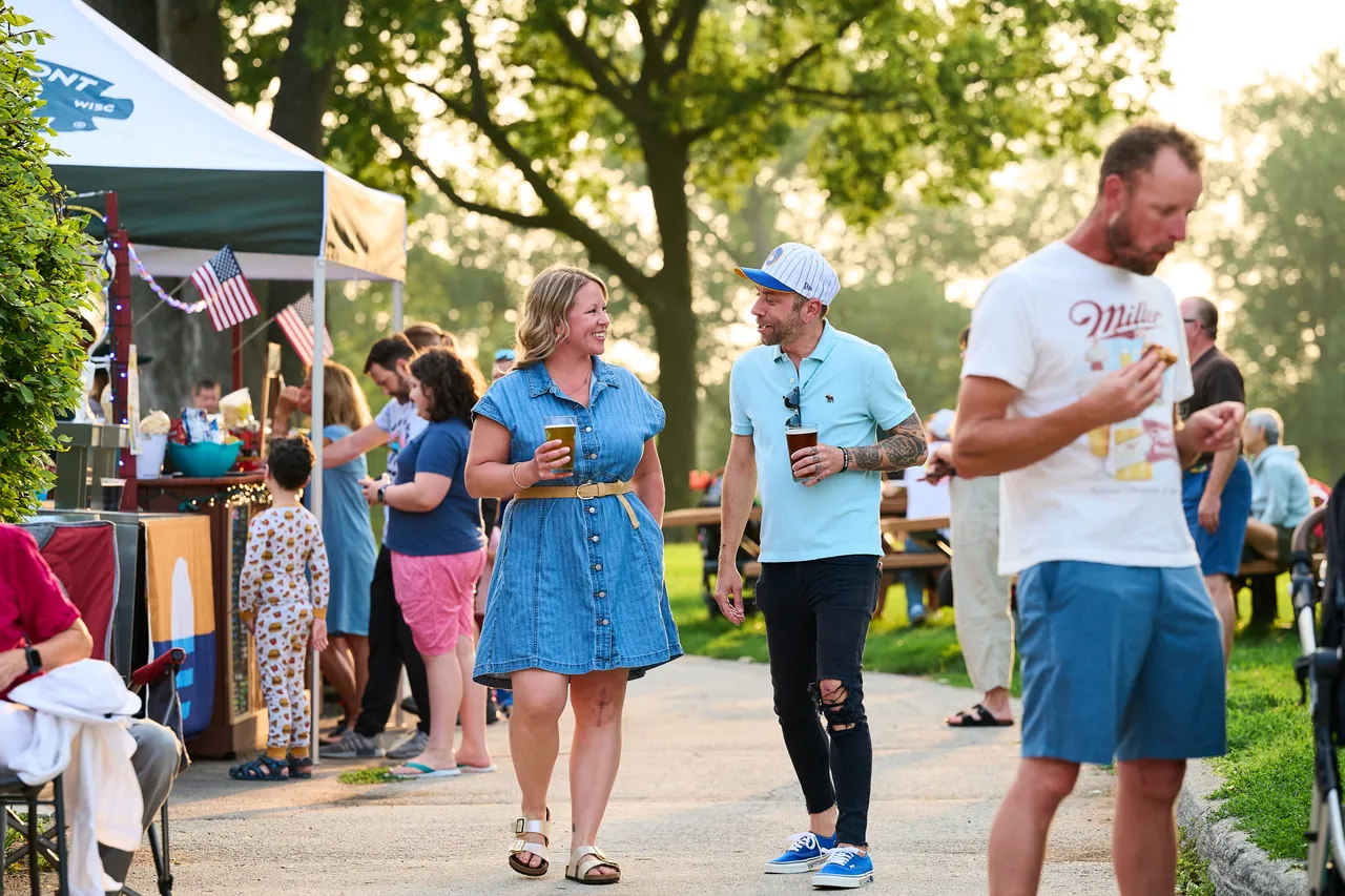 Friends chat with drinks at a sunny outdoor festival beside a vendor tent.