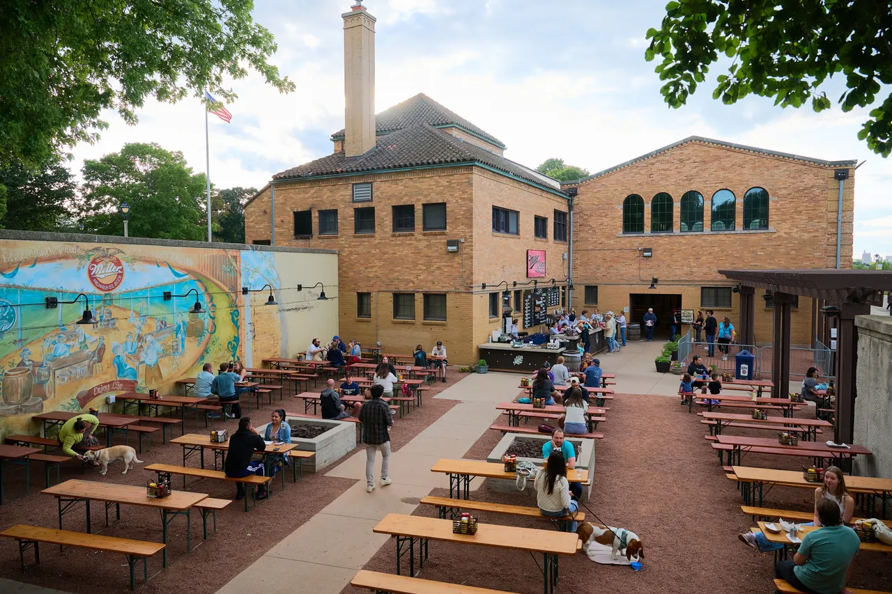 People relax at picnic tables in a brick courtyard beside a colorful mural.