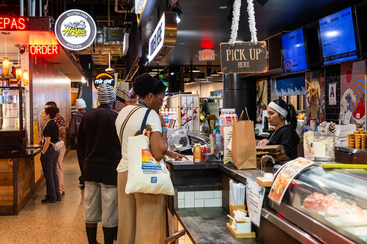 Customers order food at a busy market counter beneath neon signs and digital menu boards.
