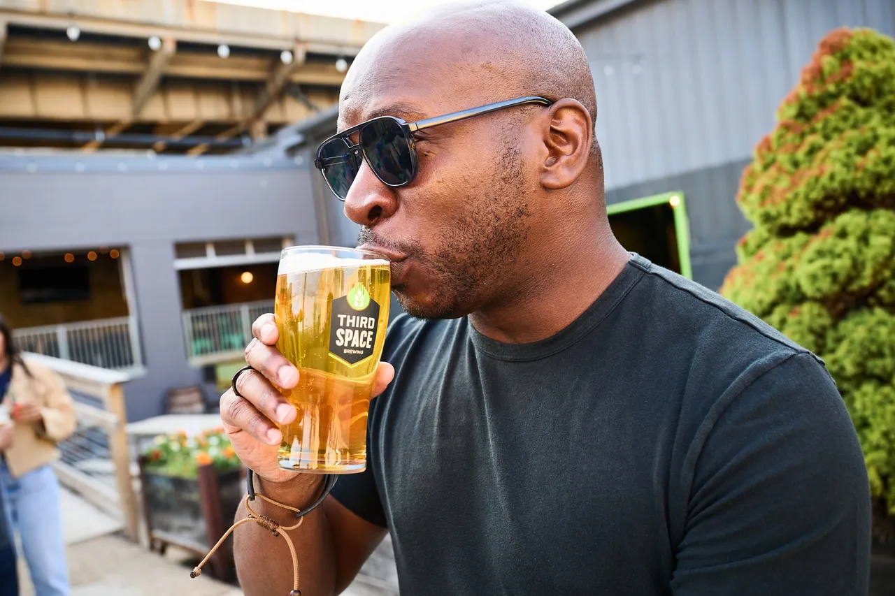 A man wearing sunglasses drinks golden beer from a Third Space Brewing glass on an outdoor patio.