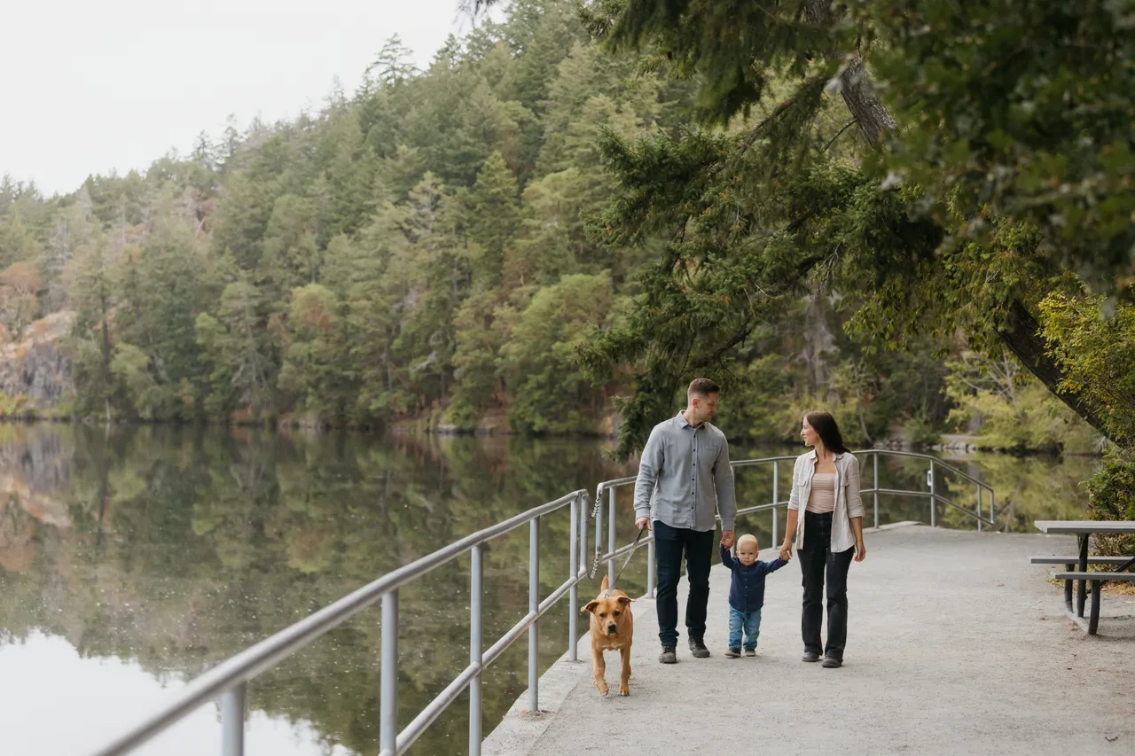 Couple walking dog near tranquil water surrounded by trees under a bright sky