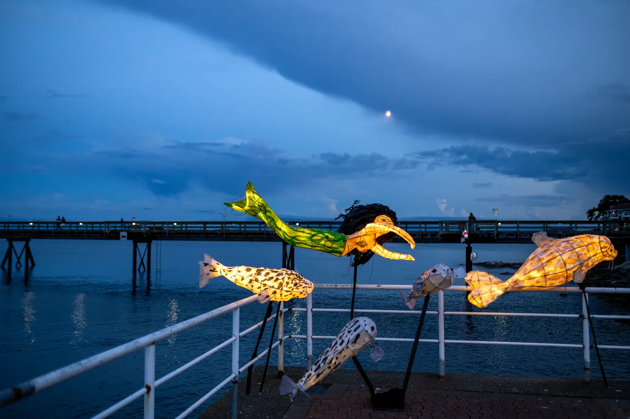 Fish and mermaid lanterns mounted along a white pier railing under heavy evening clouds
