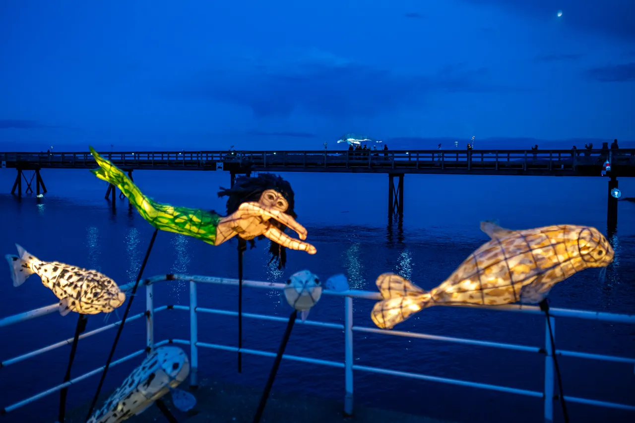 Fish and mermaid lanterns glowing against a deep blue twilight sky over the water