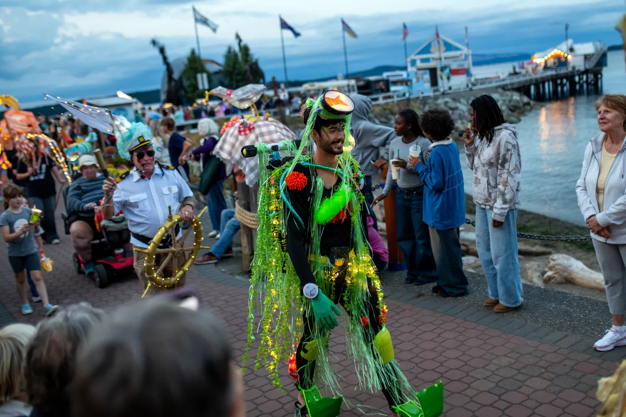 Performer in Green Seaweed Costume Walks Waterfront Promenade During Lantern Parade