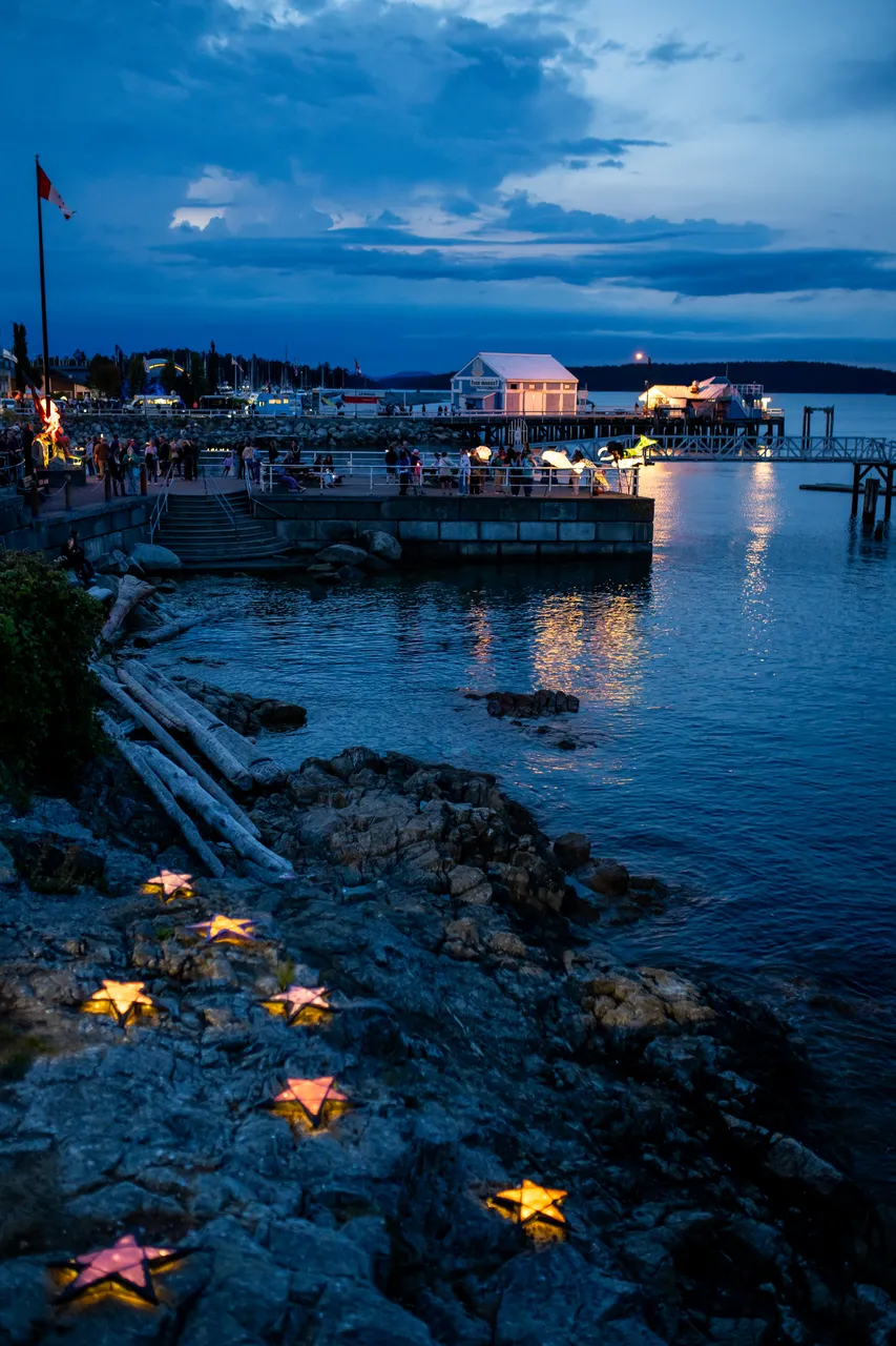Small glowing lanterns scattered across a rocky shore with a marina beyond