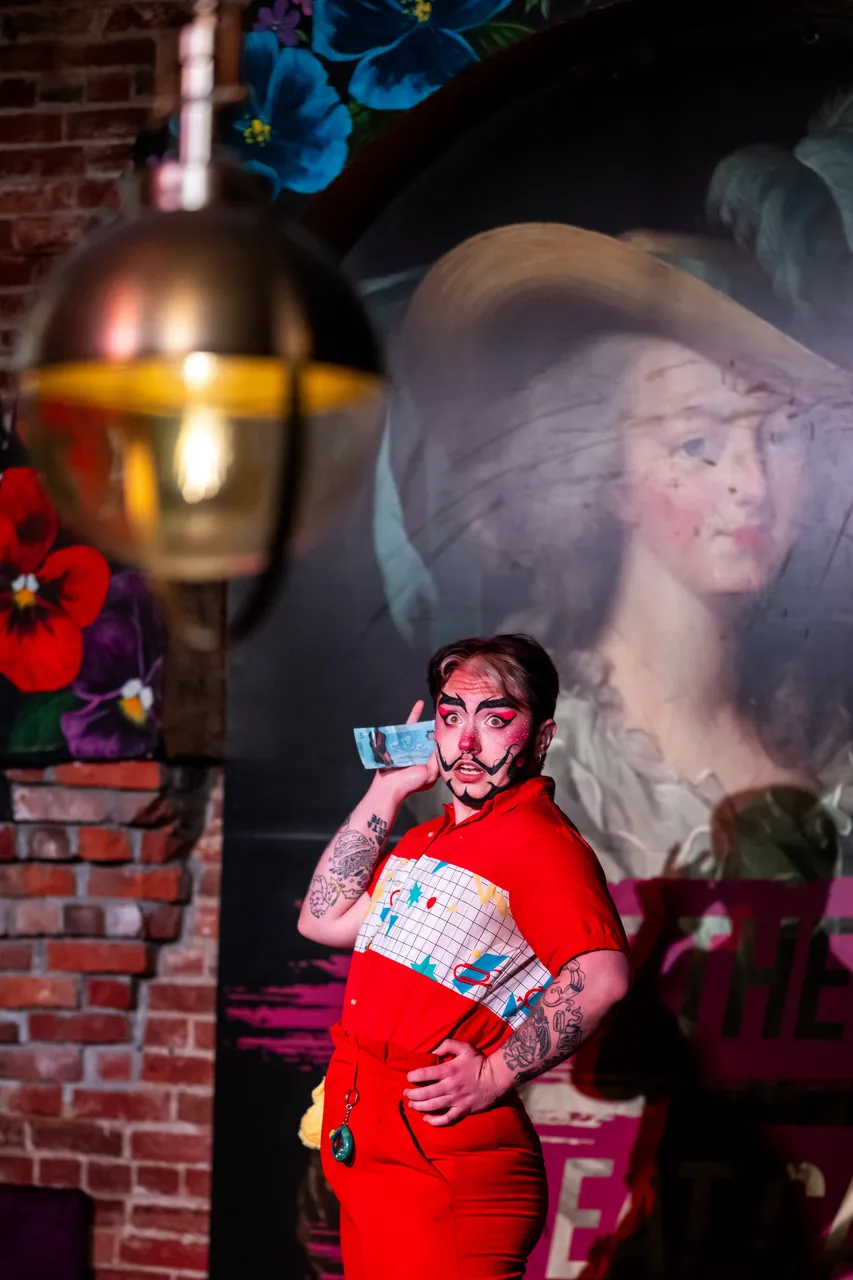 Performer Holding Money to Ear with Painting of Woman in Hat in Background