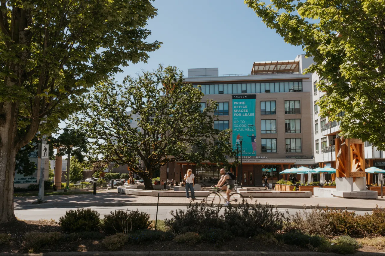 Man Biking Past Greater Victoria Public Library Surrounded by Trees on a Sunny Day