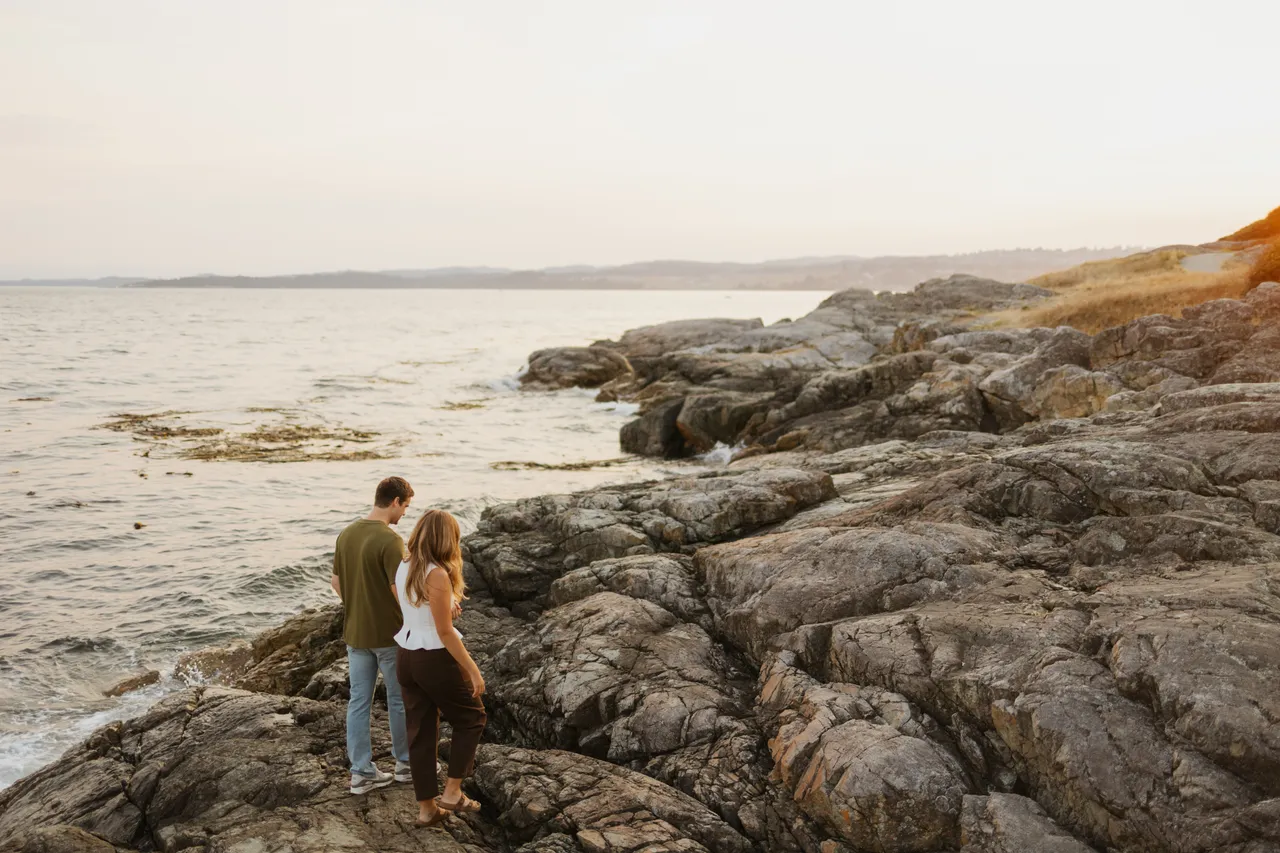 Couple stand on golden evening light over a rocky coastal area near water
