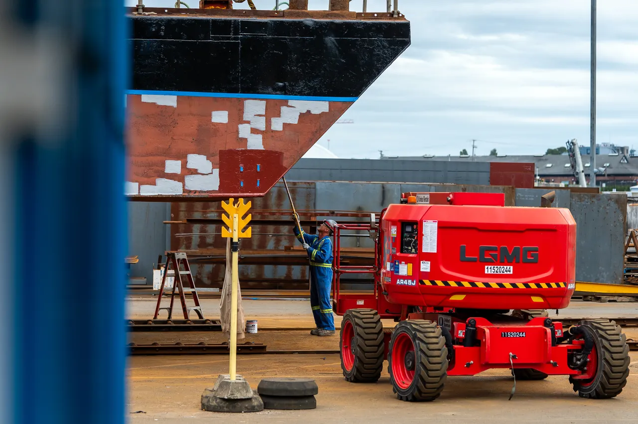 Shipyard Worker Painting a Ship Hull Beside a Red Boom Lift