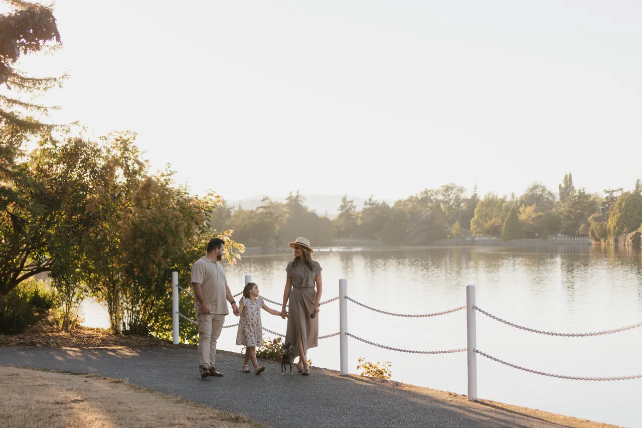 Family of three walks along body of water with trees on sunny day