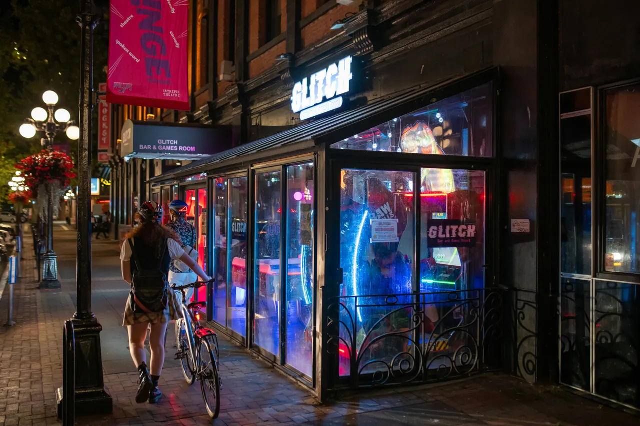 Cyclist passes brightly lit storefronts and neon signage on a city street at night