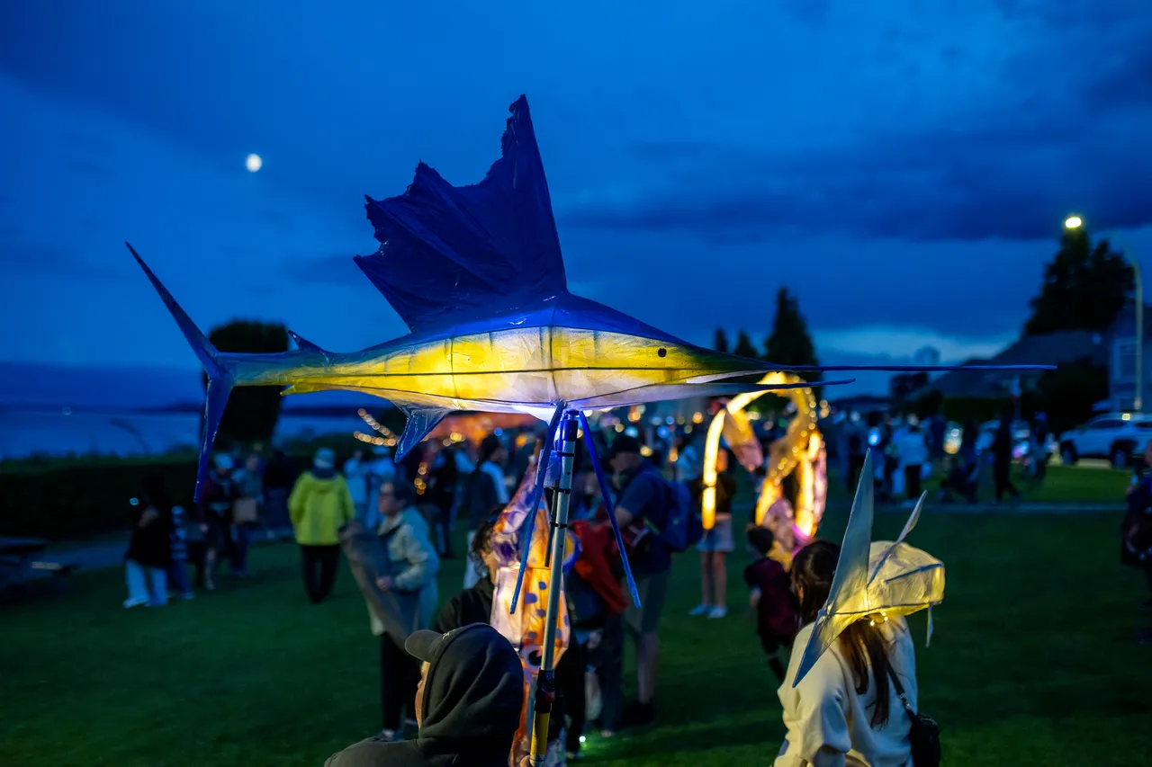 Blue-finned sailfish lantern carried above a crowd gathered on a waterfront lawn