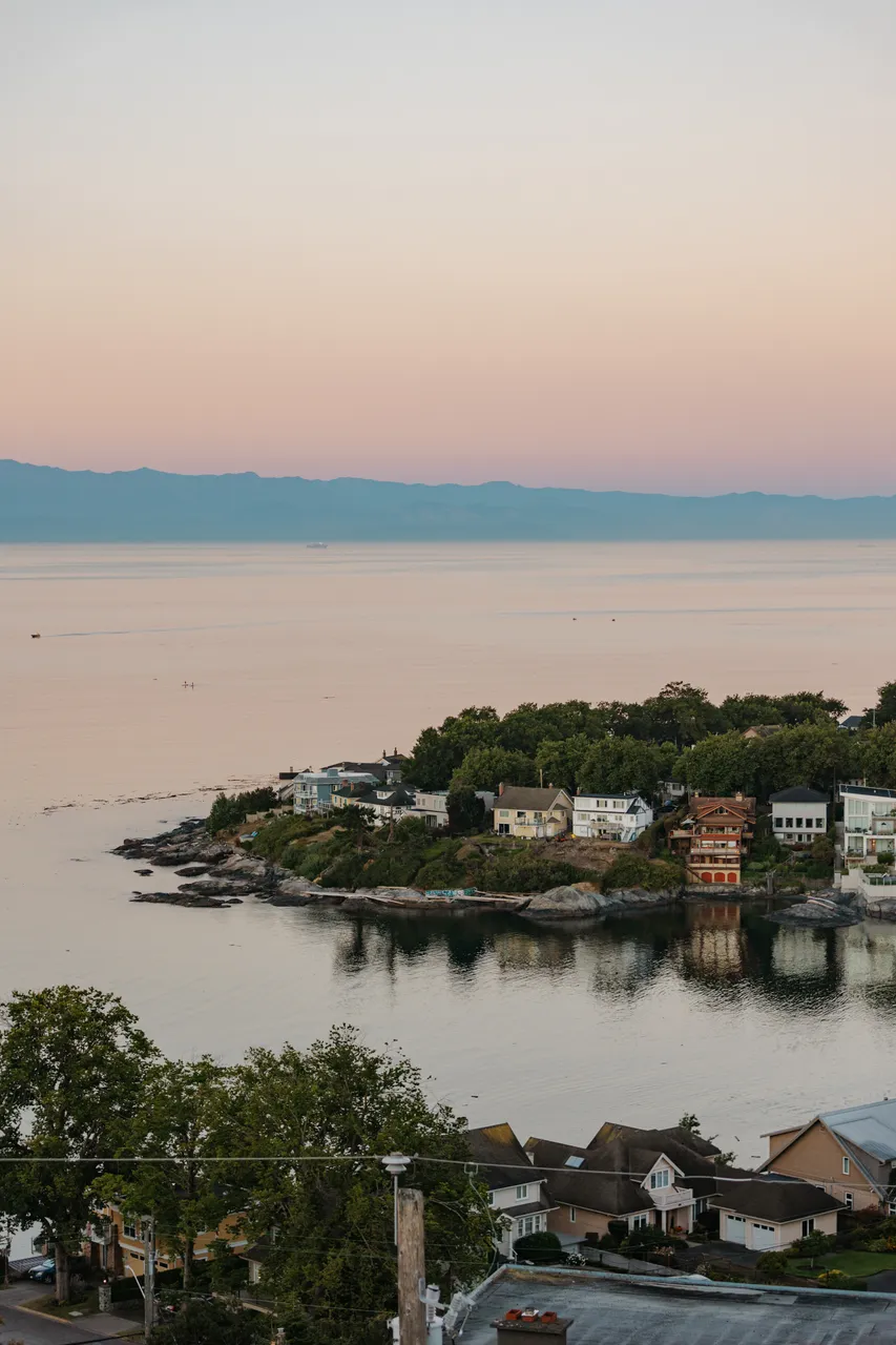 Coastal houses on a rocky outcrop surrounded by calm water with mountains in the distance.