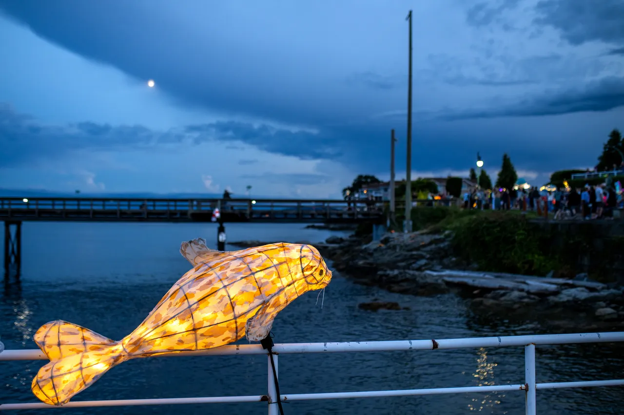 Large illuminated fish lantern resting on a rocky shoreline beside a pier at dusk
